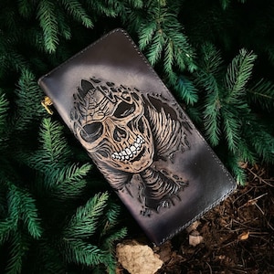 Pode incluir: Carteira de couro preta com um design detalhado de caveira. A caveira é intrincadamente esculpida e colorida com detalhes em castanho e branco. A carteira tem um zíper dourado e está sobre agulhas de pinheiro verdes.