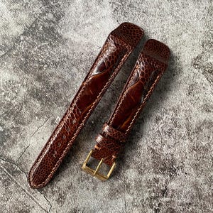 Puede incluir: Dos correas de reloj de cuero marrón con textura de piel de reptil. Una correa tiene una hebilla dorada. Las correas están en ángulo sobre una superficie gris texturizada. Las costuras son visibles a lo largo de los bordes.