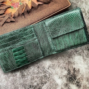 May include: A green leather wallet with a textured pattern, featuring multiple card slots and a bill compartment. The wallet is open, displaying its interior. The background includes a brown leather-bound notebook with floral accents.