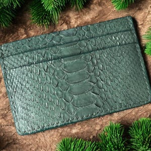 May include: A teal green cardholder with a snakeskin texture. The rectangular wallet has multiple card slots and is surrounded by green pine branches. The cardholder is made of a durable material.