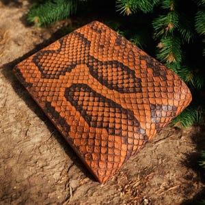 May include: A rectangular, orange and black snakeskin-patterned wallet. The wallet is made of textured leather and is set against a natural, earthy background with green foliage.