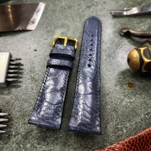 May include: A pair of blue leather watch straps with a gold buckle, alongside leatherworking tools. The straps have a textured pattern and are designed for a watch. The tools include a blade, punches, and a burnishing tool, suggesting a leathercraft theme.