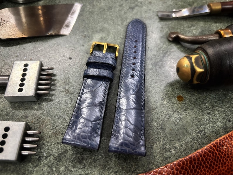 May include: A pair of blue leather watch straps with a gold buckle, alongside leatherworking tools. The straps have a textured pattern and are designed for a watch. The tools include a blade, punches, and a burnishing tool, suggesting a leathercraft theme.