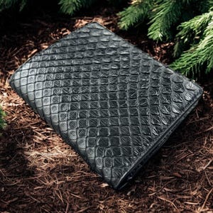 Puede incluir: Una cartera negra con un patrón texturizado en forma de escamas. La cartera es rectangular y parece ser de cuero. La cartera está rodeada de agujas de pino y descansa sobre un lecho de mantillo marrón.