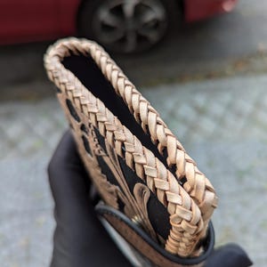 May include: A rectangular leather case featuring a black central panel with intricate tan floral tooling. The edges are finished with a thick, hand-braided tan leather lacing that creates a textured border along the top and sides of the item.