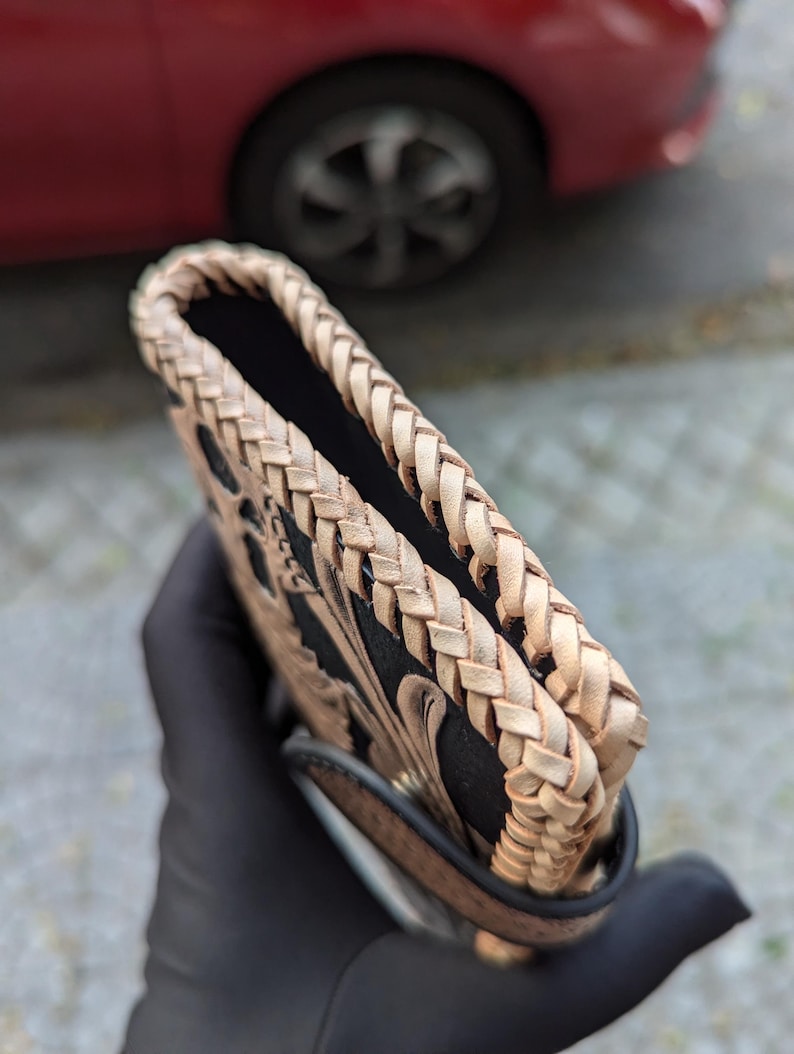 May include: A rectangular leather case featuring a black central panel with intricate tan floral tooling. The edges are finished with a thick, hand-braided tan leather lacing that creates a textured border along the top and sides of the item.