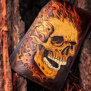 May include: A brown leather wallet featuring a detailed skull design with flames. The skull is tan, and the flames are orange and red, contrasting against the dark brown leather. The wallet is a rectangular shape.