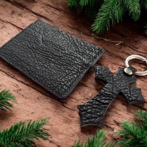 May include: A black textured leather card holder and a matching black textured leather cross-shaped keyring with a silver metal ring. Both items feature a distinct pebbled surface texture and are presented together as a set.