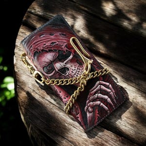 May include: A leather wallet with a detailed skull design in shades of red and black. The wallet features a gold-coloured chain and clasp. The design includes a skull with a red hood and skeletal hands.