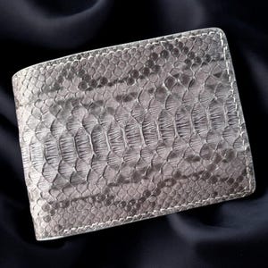 May include: A silver bi-fold wallet with a snakeskin pattern. The wallet is made of a textured material and features white stitching around the edges. The wallet is rectangular and appears to be closed. The background is a dark, solid color.