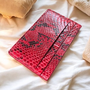 May include: A red and black snakeskin-patterned cardholder. The wallet has a rectangular shape with three visible card slots. The leather accessory is lying on a white surface.