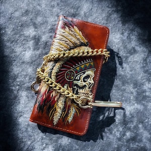 Op de afbeelding: Bruin leren portemonnee met een schedel die een Indiaanse hoofdtooi draagt. Het heeft een gouden ketting en sluiting. Het schedelontwerp bevat rode, gele en witte veren.