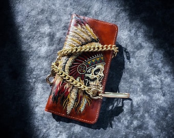 Op de afbeelding: Bruin leren portemonnee met een schedel die een Indiaanse hoofdtooi draagt. Het heeft een gouden ketting en sluiting. Het schedelontwerp bevat rode, gele en witte veren.