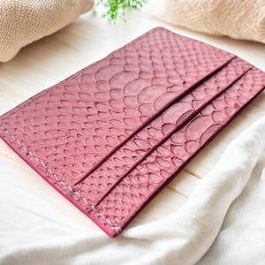 May include: A pink leather cardholder with a snakeskin pattern. The cardholder has three card slots and is stitched around the edges. The accessory is made of a textured material and is displayed on a white wooden surface.