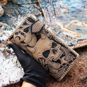 May include: A rectangular leather wallet featuring intricate hand-tooled skull motifs in tan and black. The wallet has a braided leather edge, a textured snap-button closure, and a detailed embossed surface with multiple skull and horn designs.