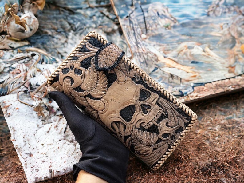May include: A rectangular leather wallet featuring intricate hand-tooled skull motifs in tan and black. The wallet has a braided leather edge, a textured snap-button closure, and a detailed embossed surface with multiple skull and horn designs.