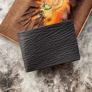 Puede incluir: Una cartera de cuero negro con una superficie texturizada. La cartera es rectangular y parece ser plegable. Descansa sobre una superficie marrón texturizada con un diseño decorativo.