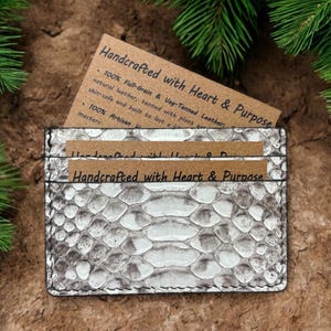Op de afbeelding: Een rechthoekige kaarthouder met een gestructureerd, wit en grijs slangenleerpatroon. Het item bevat twee bruine papieren inzetstukken met de gedrukte tekst Handcrafted with Heart & Purpose, met materiaalinformatie in zwarte inkt.
