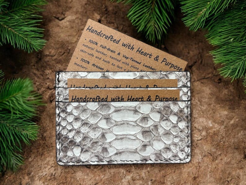 Op de afbeelding: Een rechthoekige kaarthouder met een gestructureerd, wit en grijs slangenleerpatroon. Het item bevat twee bruine papieren inzetstukken met de gedrukte tekst Handcrafted with Heart & Purpose, met materiaalinformatie in zwarte inkt.