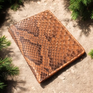 May include: A rectangular, brown leather wallet with a snakeskin pattern. The wallet is a warm brown colour with darker brown markings. The texture of the leather is visible. The wallet is surrounded by green pine needles.
