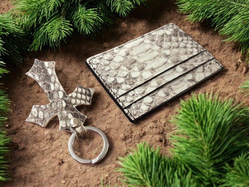 Op de afbeelding: Een rechthoekige kaarthouder en een kruisvormige sleutelhanger, beide gemaakt van materiaal met een grijs en wit slangenleermotief. De kaarthouder heeft meerdere vakjes en de sleutelhanger is voorzien van een zilverkleurige metalen ring.