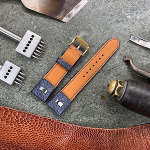 May include: A close-up of leather crafting tools and materials. Two-tone watch straps in tan and navy blue with a gold buckle are next to various metal tools. A piece of brown textured leather is in the foreground.