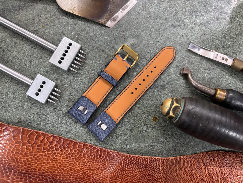 May include: A close-up of leather crafting tools and materials. Two-tone watch straps in tan and navy blue with a gold buckle are next to various metal tools. A piece of brown textured leather is in the foreground.