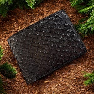 May include: A black, textured wallet with a scale-like pattern, resting on a bed of brown mulch and surrounded by green pine branches. The wallet appears to be made of leather.