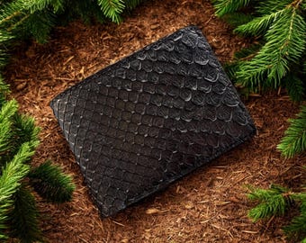 Peut inclure: Un portefeuille noir texturé avec un motif en écailles, posé sur un lit de paillis brun et entouré de branches de pin vert. Le portefeuille semble être en cuir.