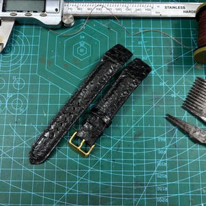 May include: A black textured leather watch strap with a gold-toned metal buckle. The strap is positioned near a stainless steel digital caliper, a spool of copper wire, and metal leatherworking tools on a green cutting mat with grid lines.