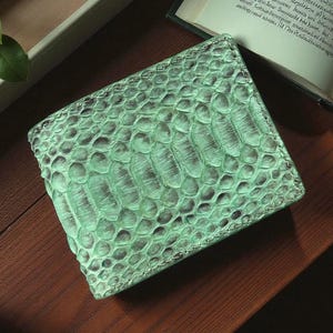 May include: A bi-fold wallet with a textured, snake-skin pattern in shades of mint green and brown. The wallet is rectangular and appears to be made of leather. The wallet is on a wooden surface, with a book and leaves in the background.