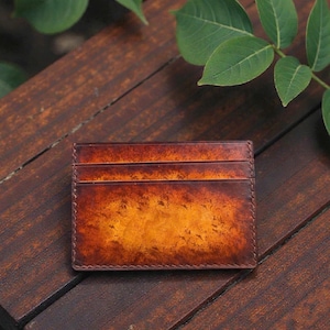 May include: A brown leather cardholder with a gradient effect, shifting from dark brown to golden-orange. It features multiple card slots and stitched edges. The background is a wooden surface with green leaves. The cardholder is approximately 10 cm in length.