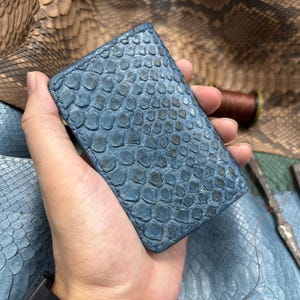 May include: A rectangular card holder crafted from blue snakeskin leather with a distinct scale texture. The edges are stitched with matching blue thread, showcasing a natural pattern of overlapping scales across the entire surface.