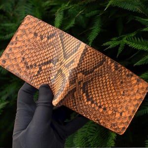 May include: A rectangular wallet with an orange and black snakeskin pattern. The wallet is displayed against a backdrop of green foliage, highlighting its textured surface and design.