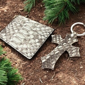 Op de afbeelding: Een rechthoekige kaarthouder en een kruisvormige sleutelhanger, beide met een gestructureerd grijs en wit slangenleerpatroon. De sleutelhanger bevat een ronde zilverkleurige metalen ring die met een kleine zilveren klinknagel is bevestigd.
