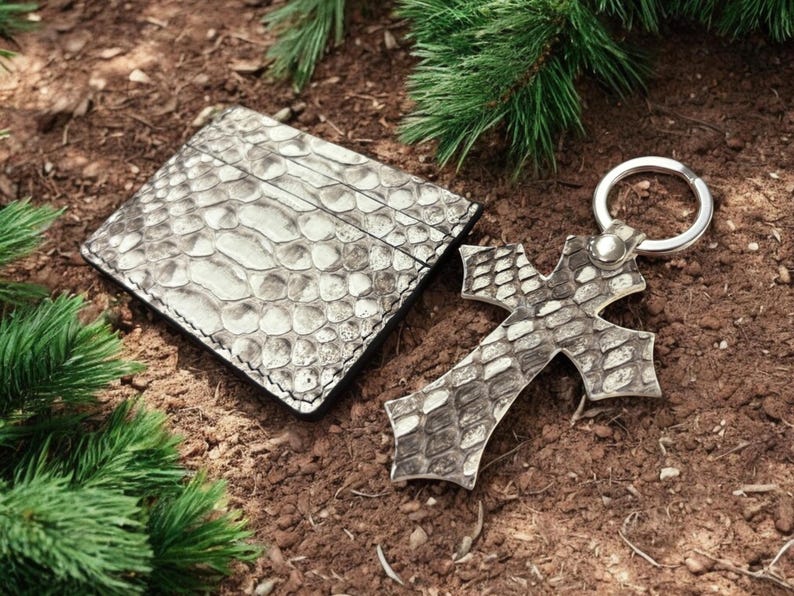 Op de afbeelding: Een rechthoekige kaarthouder en een kruisvormige sleutelhanger, beide met een gestructureerd grijs en wit slangenleerpatroon. De sleutelhanger bevat een ronde zilverkleurige metalen ring die met een kleine zilveren klinknagel is bevestigd.