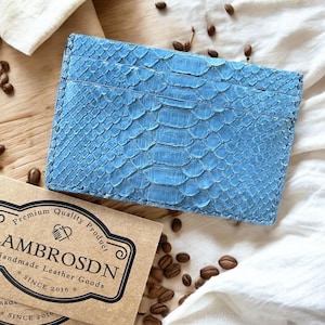 Peut inclure: Un porte-cartes en cuir bleu clair avec un motif peau de serpent. Le porte-cartes est rectangulaire avec plusieurs fentes pour cartes. La marque "LAMBROSDN" est imprimée sur une carte marron. Articles de maroquinerie faits à la main depuis 2016.