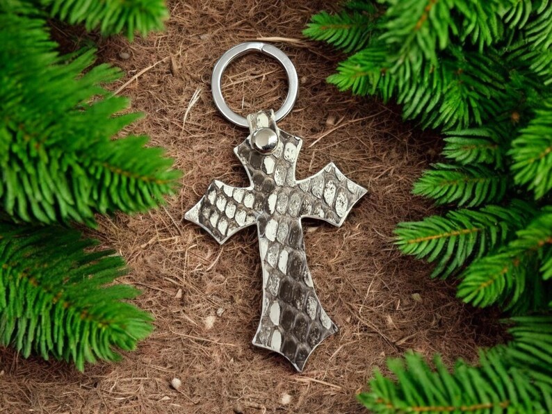 Op de afbeelding: Een kruisvormige sleutelhanger met een textuur in slangenleerpatroon in grijze, witte en zwarte tinten. Het kruis is bevestigd aan een zilverkleurige metalen ring door een klein, bijpassend leren lipje met een centrale metalen klinknagel.