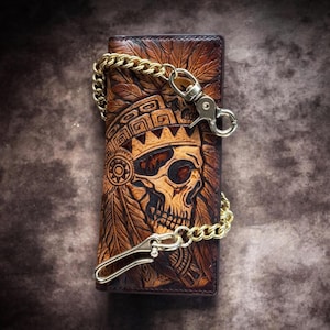 May include: A brown leather wallet with a carved skull design, featuring a gold chain and clasps. The wallet showcases detailed carvings of feathers and geometric patterns, offering a distinctive accessory.