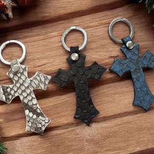 Puede incluir: Tres llaveros en forma de cruz con un patrón de piel de serpiente texturizada. Los llaveros son de color gris claro, negro sólido y azul oscuro. Cada cruz está unida a una argolla de metal plateado mediante un pequeño remache de metal.