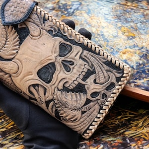 May include: A rectangular leather wallet featuring intricate hand-tooled carvings of two skulls with horns and leaf-like patterns. The design uses a contrast between natural tan leather and black dyed recessed areas, finished with a braided leather edge.