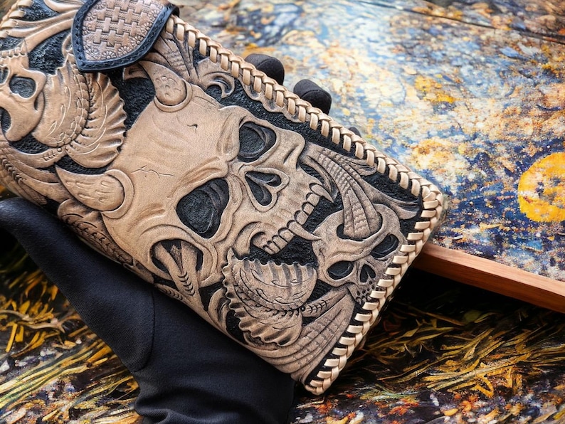 May include: A rectangular leather wallet featuring intricate hand-tooled carvings of two skulls with horns and leaf-like patterns. The design uses a contrast between natural tan leather and black dyed recessed areas, finished with a braided leather edge.
