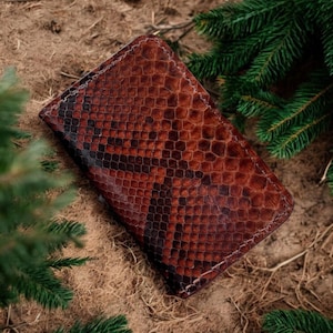 以下が含まれることがあります： 茶色と黒のヘビ柄の財布。長方形の財布にはステッチの縁取りがあります。背景には茶色のマルチと緑の松の枝があります。スタイリッシュなアクセサリーです。