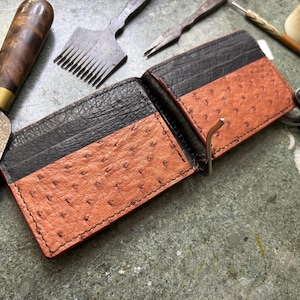 May include: A bi-fold wallet featuring a combination of black textured leather and tan ostrich-embossed leather. The wallet includes a silver-toned metal money clip attached to the centre fold, with visible black stitching along the edges.