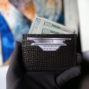 Puede incluir: Un tarjetero de cuero negro texturizado con dos billetes de 100 euros y varias tarjetas. El tarjetero se sostiene en una mano enguantada de negro. El tarjetero tiene un diseño minimalista.