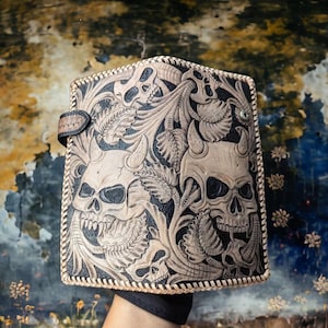 May include: A rectangular leather wallet featuring intricate hand-tooled carvings of multiple horned skulls integrated with detailed floral and leaf patterns. The edges are finished with a decorative whipstitch lacing and a small side snap closure.