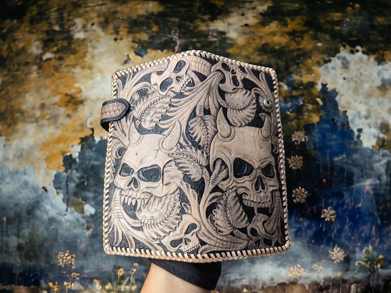 May include: A rectangular leather wallet featuring intricate hand-tooled carvings of multiple horned skulls integrated with detailed floral and leaf patterns. The edges are finished with a decorative whipstitch lacing and a small side snap closure.