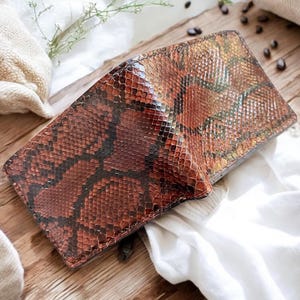 Peut inclure: Portefeuille à deux volets avec un motif peau de serpent marron et noir. Le portefeuille en cuir présente un aspect texturé. Le portefeuille est ouvert, révélant l'intérieur. Le fond comprend une surface en bois et du tissu blanc. Environ 12 cm de large.