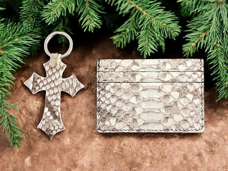 Op de afbeelding: Een kruisvormige sleutelhanger en een rechthoekige kaarthouder, beide met een gestructureerd, lichtgekleurd slangenleerpatroon. De sleutelhanger bevat een zilverkleurige metalen ring aan de bovenkant, terwijl de kaarthouder zichtbare stiksels heeft.