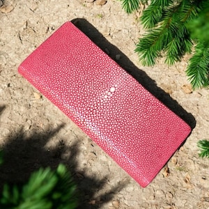 Puede incluir: Una cartera rectangular roja con una superficie texturizada, con un diseño circular blanco central. La cartera se encuentra sobre un fondo de elementos naturales, incluyendo vegetación y una superficie texturizada de color marrón claro.